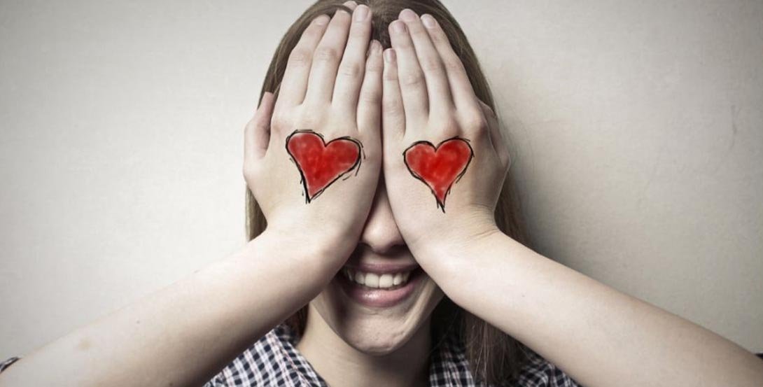 Una chica se cubre los ojos con las manos mientras sonríe. En el dorso de cada mano tiene pintado un corazón rojo.